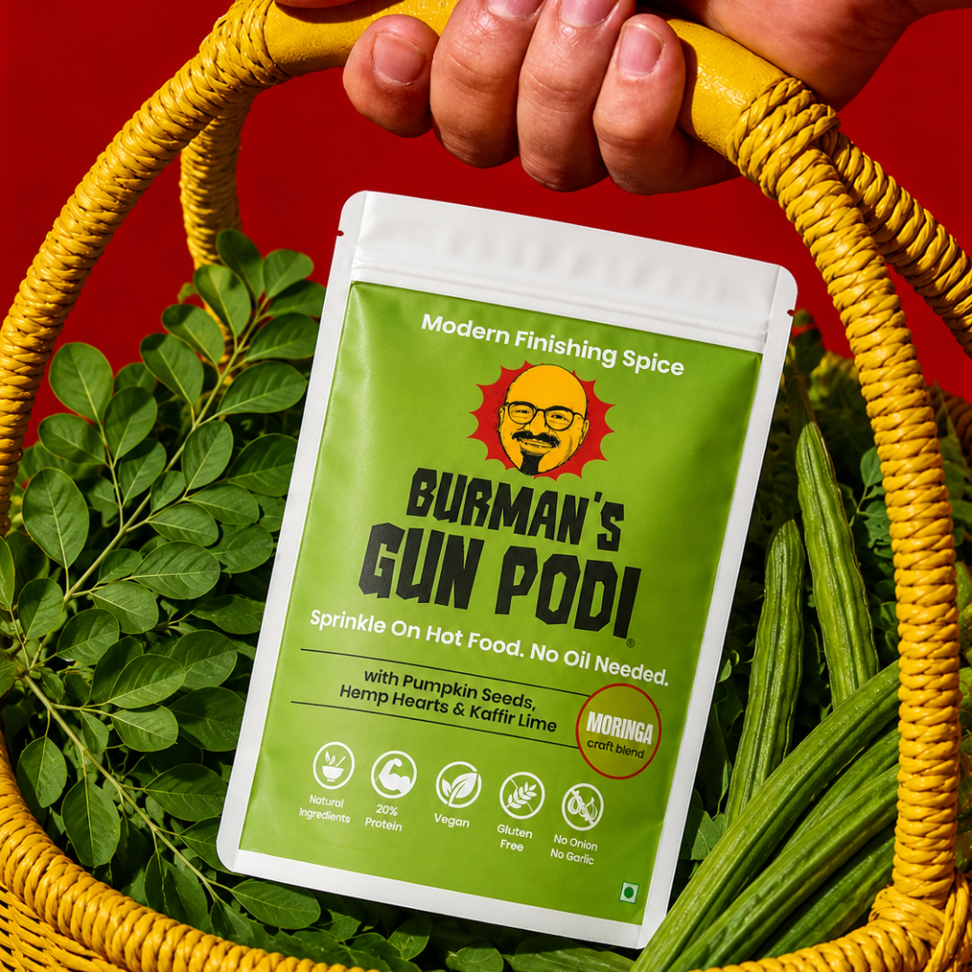 Burman's Gun Podi Moringa craft blend pouch nestled in a yellow wicker basket with fresh moringa leaves and drumsticks, highlighting its natural superfood ingredients — vegan, gluten-free, 20% protein finishing spice