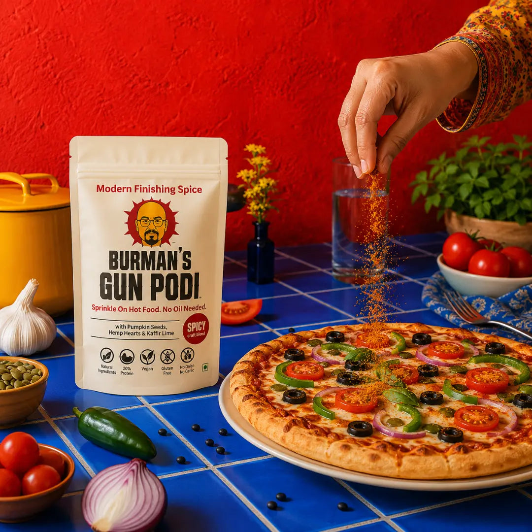 Hand sprinkling Burman's Gun Podi finishing spice over a fresh pizza topped with tomatoes, olives, and peppers - no oil needed, just sprinkle on hot food for bold flavor